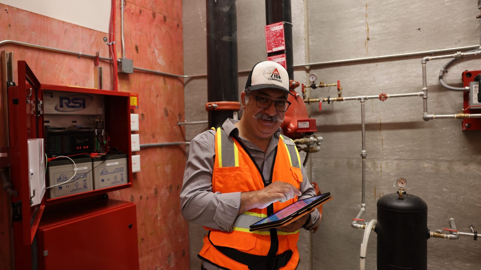 Zion technician working at a fire riser room with RSI BDA enclosure visible — sprinkler piping and gauges in background